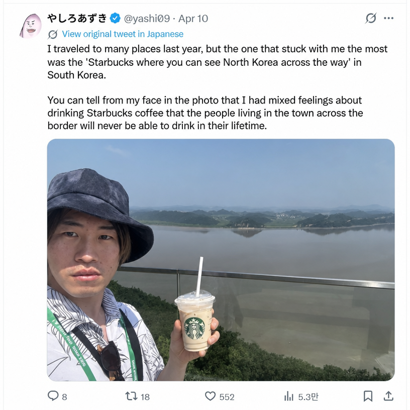 A screenshot of a tweet by Japanese manga artist Yashiro Azuki, showing a selfie holding a Starbucks coffee with a North Korean landscape in the background, making a complex, subtle expression.