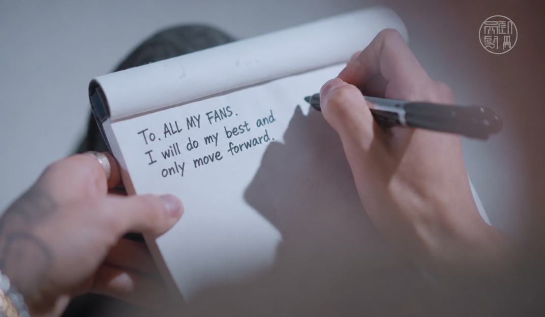 Close-up of a hand writing 'To. ALL MY FANS. I won't cause any trouble, I'll just serve my time' with a pen on a notepad.