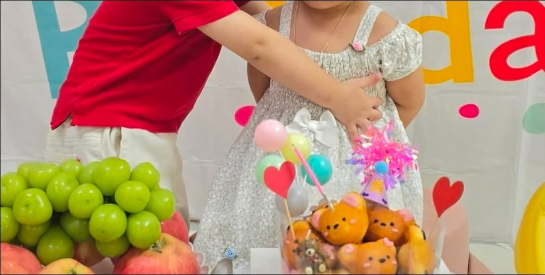 At a daycare birthday party, a boy in a red shirt approaches a girl wearing a dress, appearing ready to hug or kiss her.