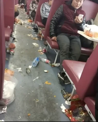 乗客がスマートフォンを見ながら座っている中、列車の通路が捨てられたゴミ、空き瓶、食べ残し、ビニール袋で完全に埋め尽くされている。