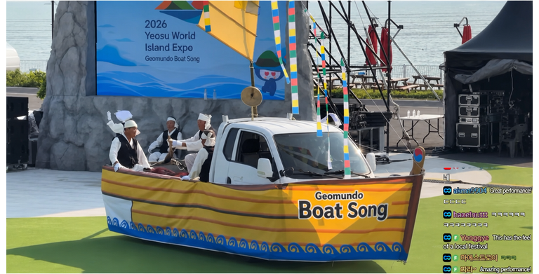 Performers on a grassy outdoor stage riding a 1-ton cargo truck decorated to look like a boat, performing the Geomundo boat song.