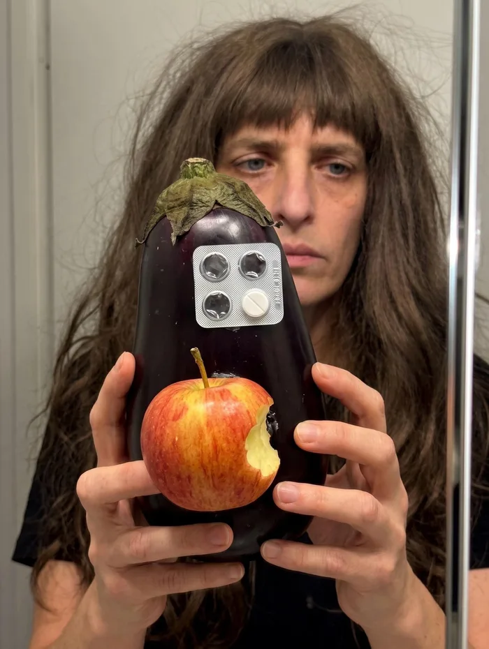 鏡越しに自撮りをする女性。手にはカメラレンズモジュールに見立てた錠剤シートと、Appleのロゴに見立てた一口かじられたリンゴが貼り付けられたナスを持っている。