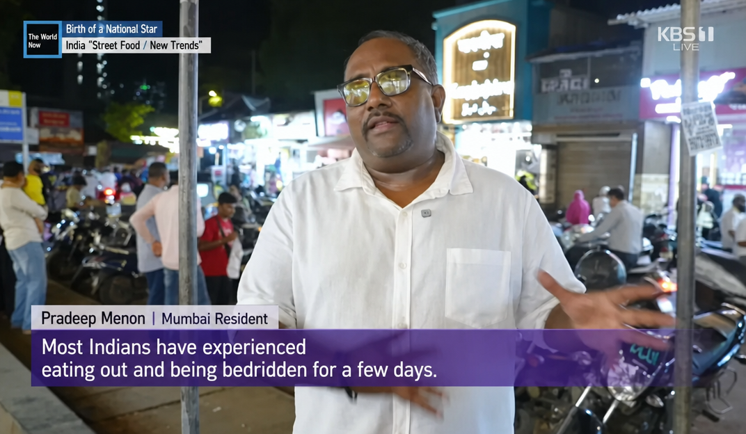 A still from a KBS1 news broadcast showing a Mumbai resident, Pradip Menon, being interviewed. He explains that most people in India have had the experience of eating out and being bedridden for several days afterwards.