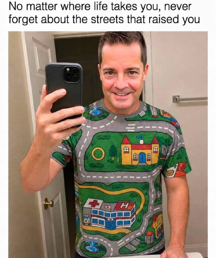 A man taking a mirror selfie wearing a t-shirt patterned like a classic kids' city street play rug, captioned: 'No matter where life takes you, never forget about the streets that raised you'.