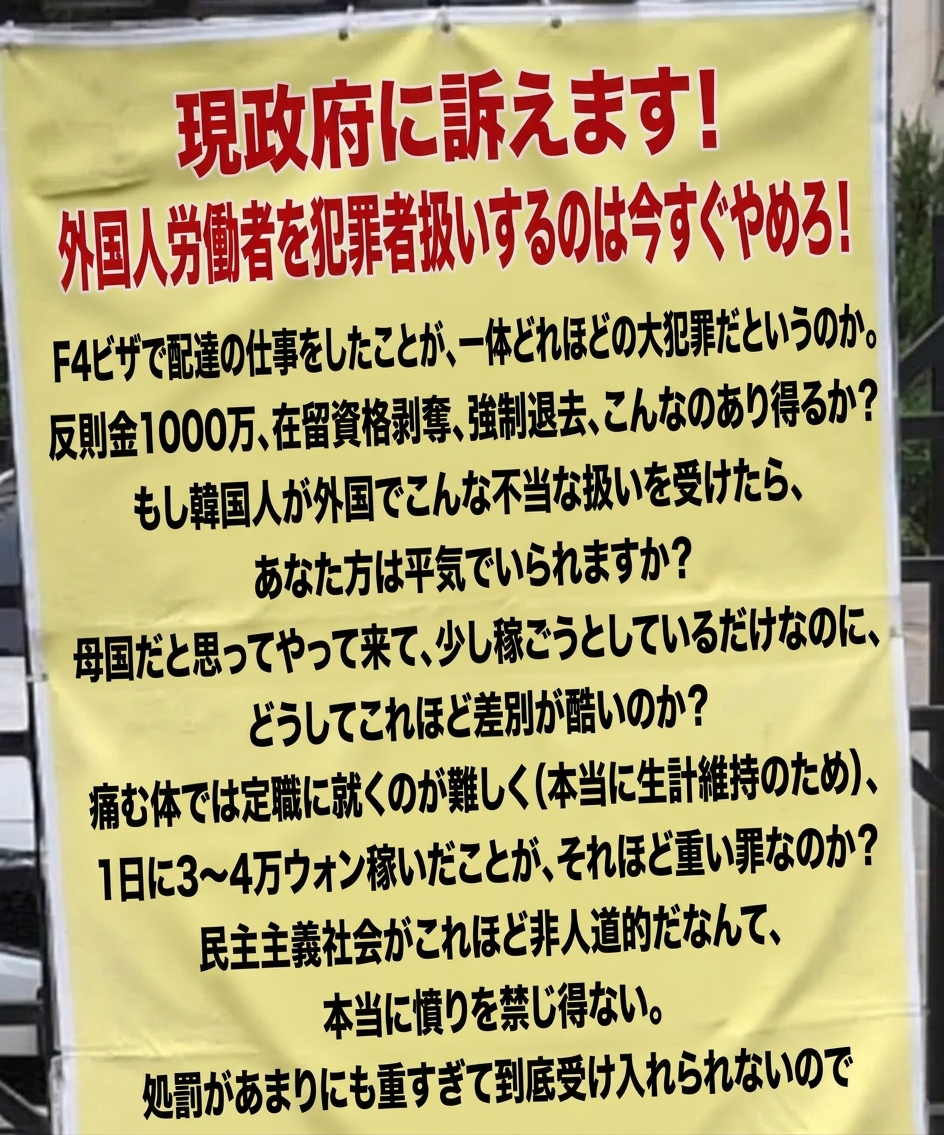 鉄柵に掛けられた黄色い横断幕。「現政府に訴えます！外国人労働者を犯罪者扱いするのは今すぐやめろ！」という見出しで、F-4ビザで配達の仕事をして摘発され、反則金1000万ウォンと強制退去処分を受けた不当さを訴える抗議文が長々と書かれている様子。