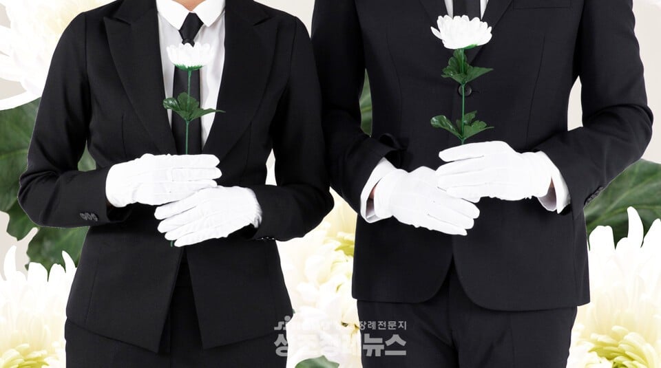 黒いスーツに身を包み、白い手袋を着けて胸の前で白い菊の花を持って立っている2人の葬儀指導員の姿。