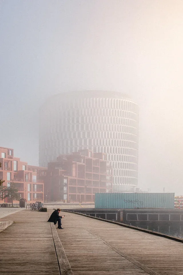 A person sitting on a wooden boardwalk near the water with heavy fog shrouding modern waterfront buildings in the background.