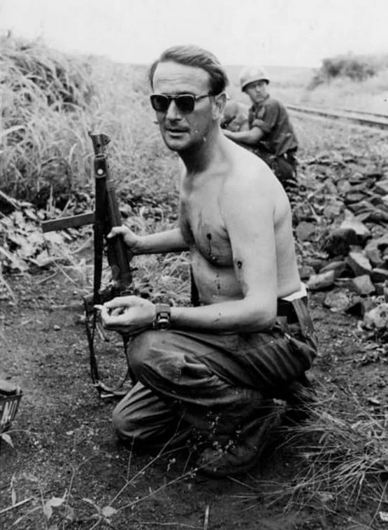 Black and white photograph showing Swedish Major Erik Bonde kneeling shirtless by railway tracks with visible bullet wounds, wearing sunglasses, holding a weapon and a cigarette.
