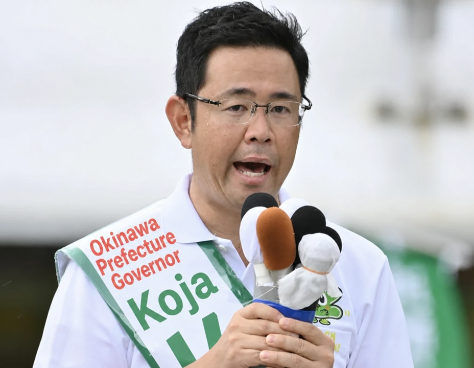 Candidate Genta Koza speaking while holding a microphone and wearing a campaign sash