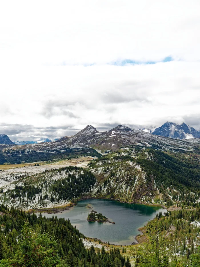 A scenic mountain landscape featuring a serene lake surrounded by evergreen forests and snowy peaks under a cloudy sky.