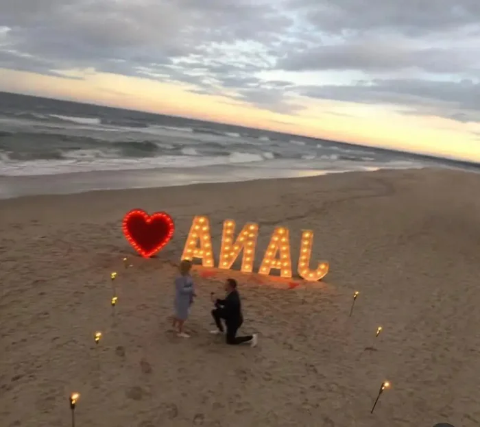 A man proposes on a beach in front of large illuminated letters spelling JANA, but viewed from behind they appear to spell ANAL next to a glowing heart.