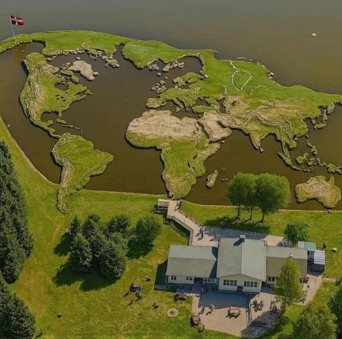 Aerial view of a landscape installation shaped like a world map on the edge of a lake next to a visitor building