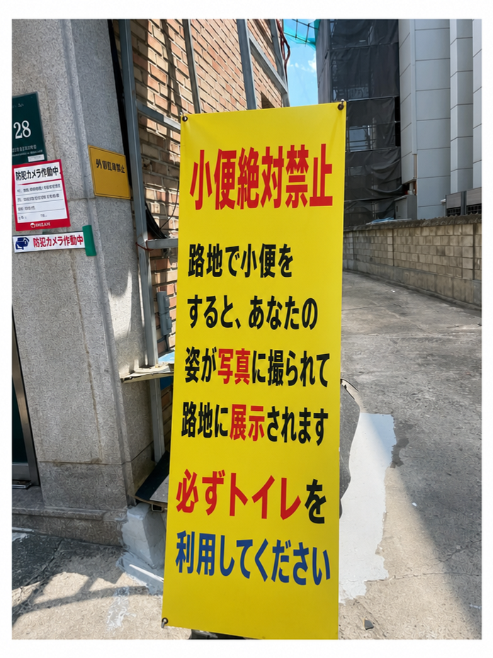 路地裏に設置された「小便絶対禁止」と書かれた黄色い警告バナー。路地で小便をすると、その姿が写真に撮られて掲示されるという内容が書かれている。