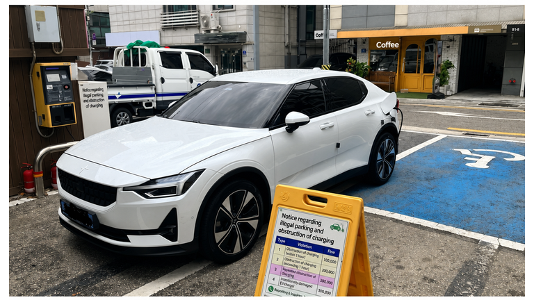 Front view of a white electric vehicle parked horizontally across parking spots and charging, behind a enforcement notice sign.