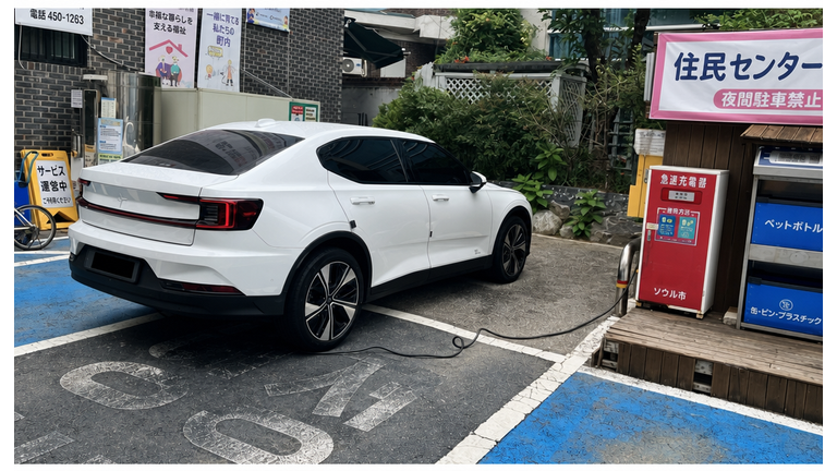 住民センターの駐車ラインをまたいで駐車し、離れた電気自動車充電器からケーブルを接続している白いポールスター車両の後部。
