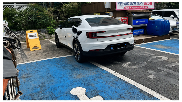 障害者専用駐車エリアのラインを踏んで横向きに駐車された白い電気自動車と、接続された充電ケーブル。
