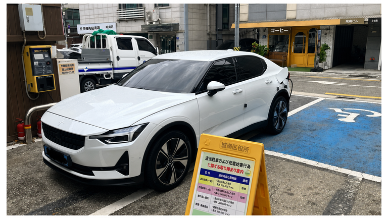 取り締まり案内板の後ろで駐車枠を無視して横向きに駐車し、充電中の白い電気自動車の正面。