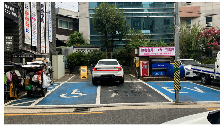 住民センターの駐車場全景。障害者用駐車エリアと一般エリアを対角線上に占拠し、充電ケーブルを引き込んでいる白い電気自動車。