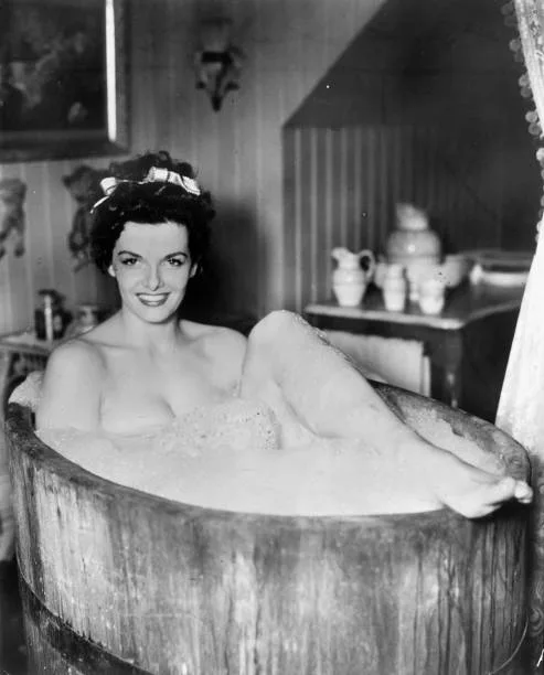 Black and white photograph of actress Jane Russell smiling while sitting in a bubble-filled rustic wooden tub with one leg resting on the rim.
