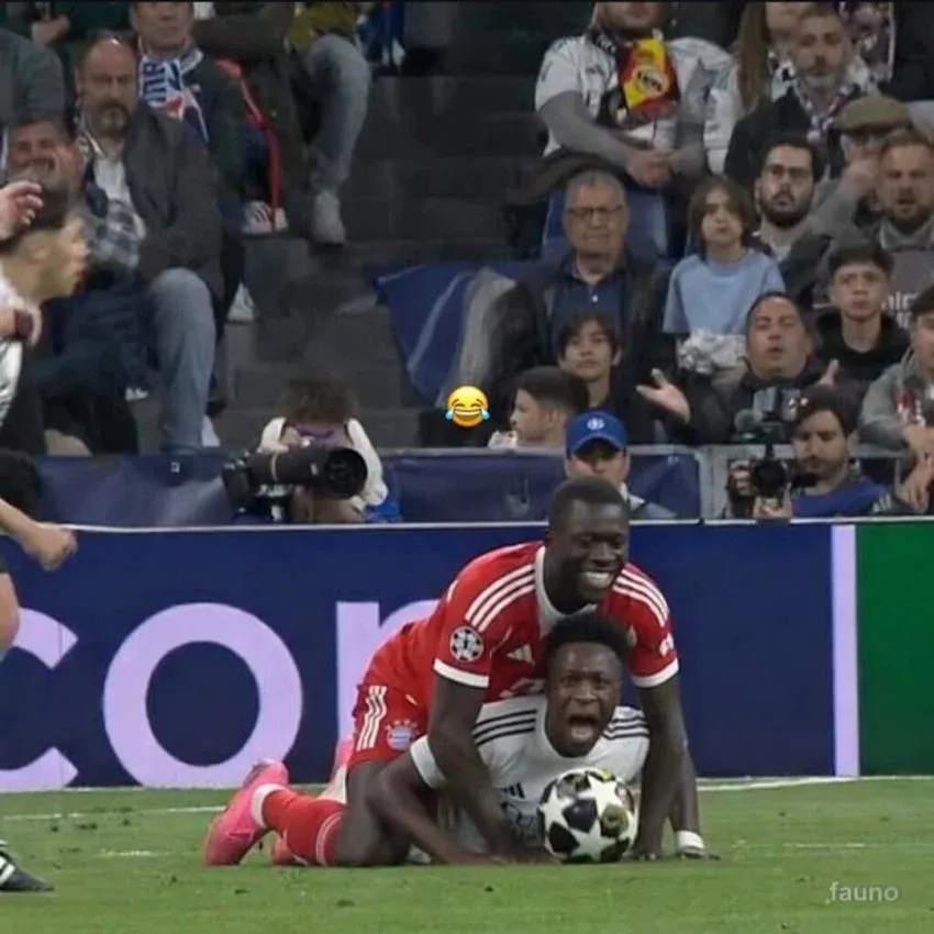 A soccer match scene where a Bayern Munich player smilingly mounts on top of a Real Madrid player who is lying face down on the ground clutching the ball and screaming.