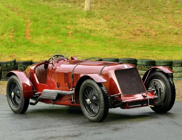 A vintage deep red 1929 Maserati Tipo V4 race car parked outdoors on asphalt in front of a grassy hill and tire barrier.