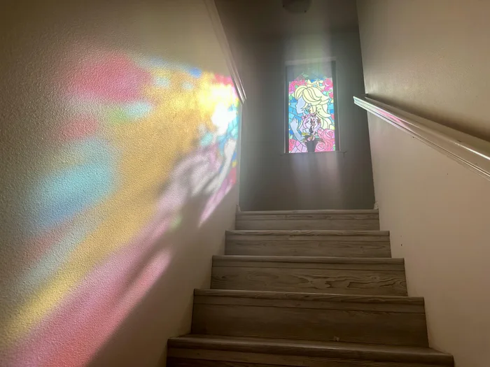 Staircase illuminated by sunlight streaming through a stained glass window depicting Princess Peach from Super Mario 64, casting colorful light patterns onto the wall.