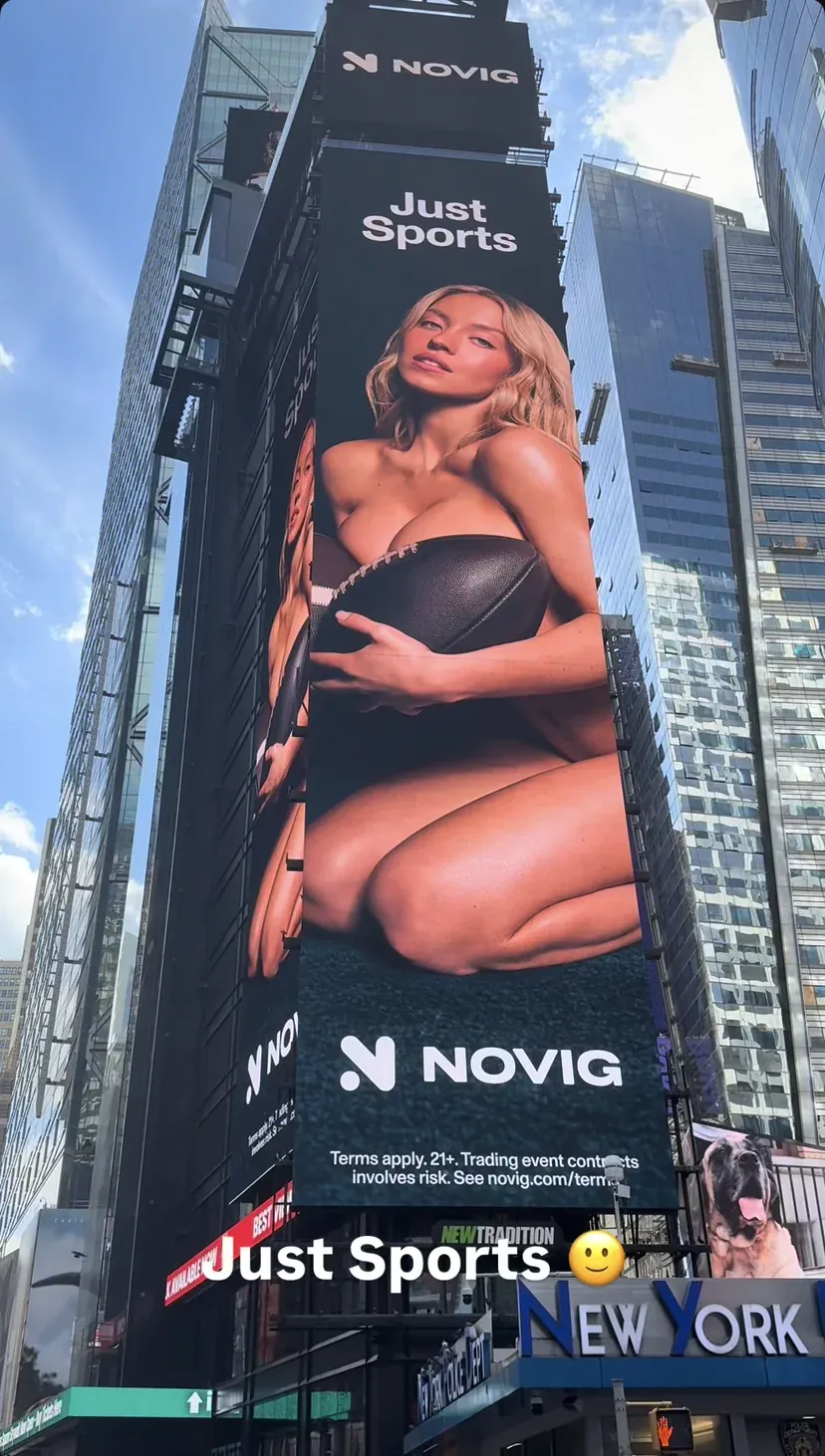 Photo of a large Novig billboard in Times Square, New York, featuring actress Sydney Sweeney topless, covering her chest with a football while kneeling.