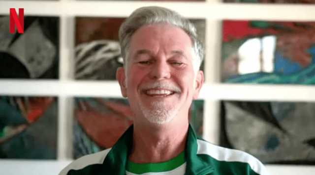 Former Netflix CEO Reed Hastings smiling broadly while wearing a green tracksuit under the Netflix logo.