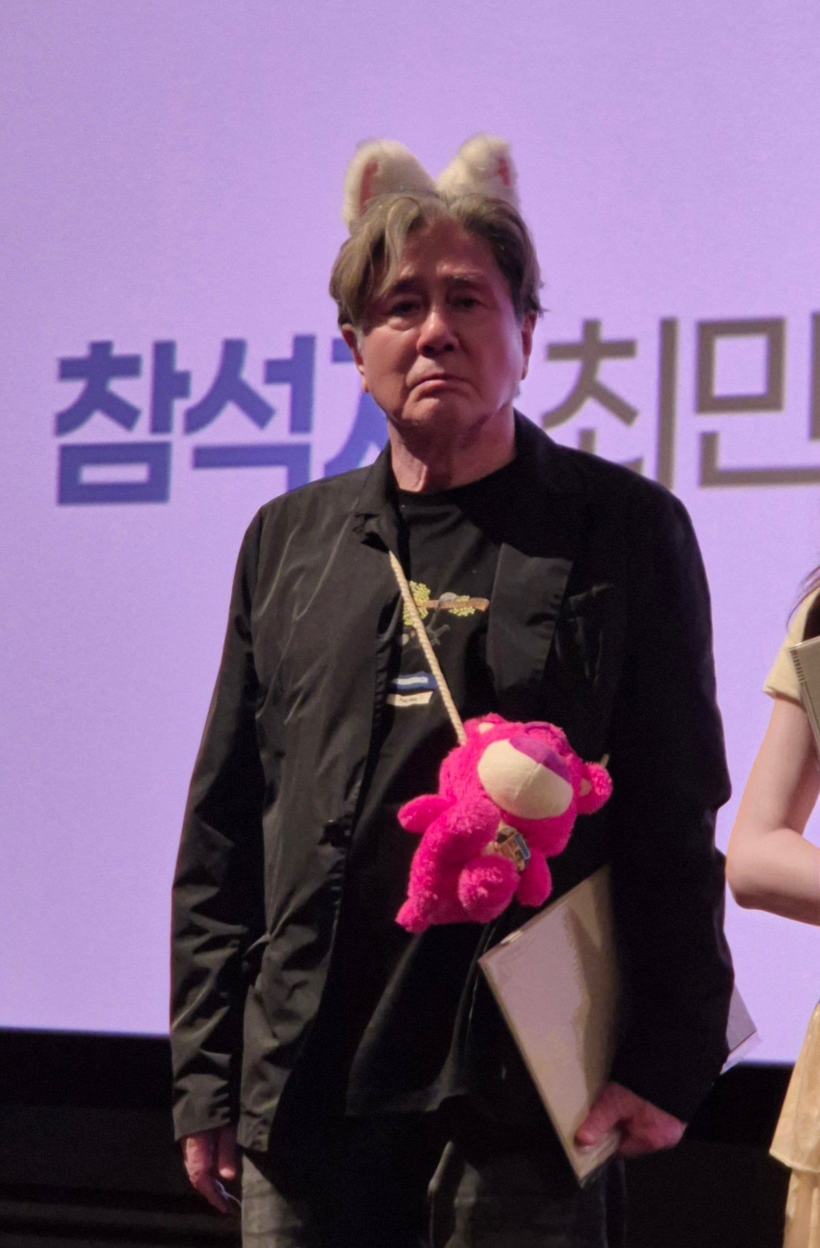 Actor Choi Min-sik making a serious expression while wearing an animal ear headband and a pink teddy bear bag