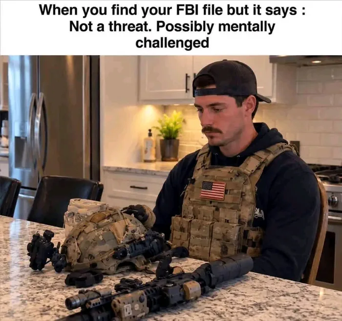 A man in tactical military gear sitting sadly at a kitchen counter looking at his helmet and rifle, with text reading: When you find your FBI file but it says: Not a threat. Possibly mentally challenged.