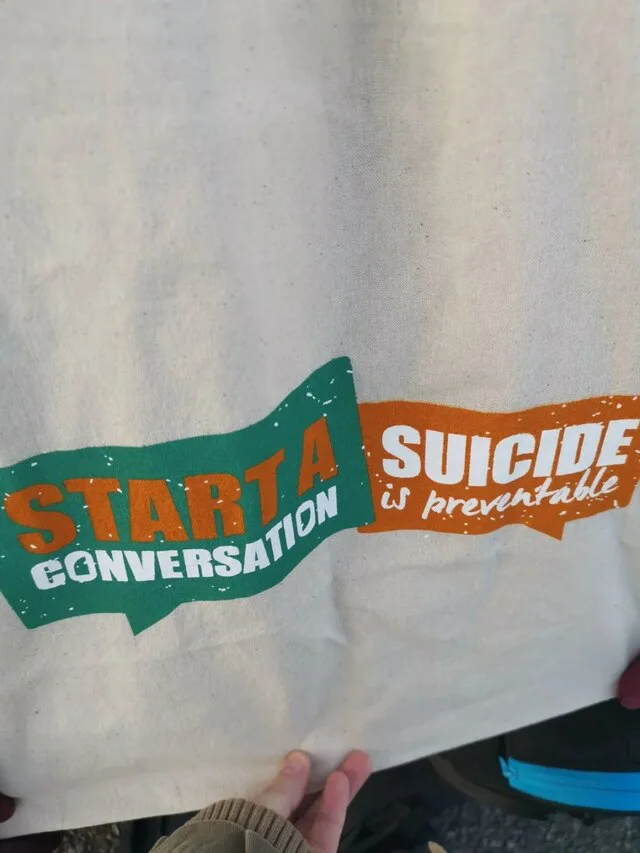 A canvas tote bag featuring two speech bubbles side-by-side with text that reads 'START A CONVERSATION' and 'SUICIDE is preventable', but the top line awkwardly reads 'START A SUICIDE'.