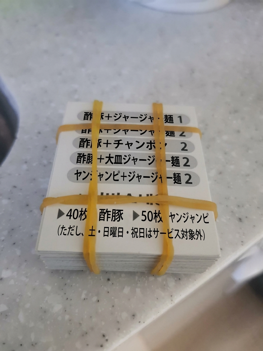 黄色い輪ゴムで束ねられた中華料理店の分厚いクーポン券の束。セットメニューの案内とともに、40枚で酢豚、50枚でヤンジャンピサービスの案内が記載されている。