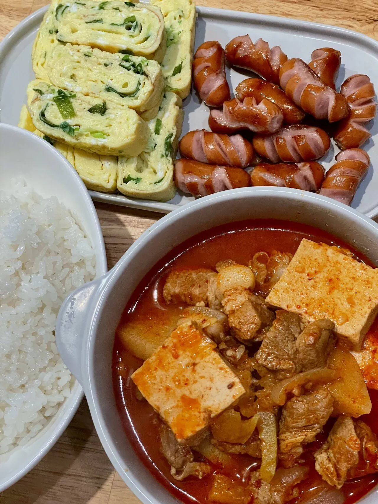 白いご飯、切り込みを入れて焼いたウィンナーソーセージ、ネギ入りの厚焼き玉子、豚肉と豆腐がたっぷり入ったキムチチゲが並べられた食卓。