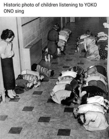 A vintage black and white photograph showing schoolchildren and teachers performing a duck-and-cover air raid drill in a school hallway, humorously captioned as children listening to Yoko Ono sing.