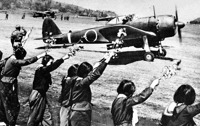 Historic black-and-white photograph from 1945 showing Japanese schoolgirls waving branches and flowers toward an Imperial Japanese Army aircraft as the pilot prepares for a kamikaze takeoff.