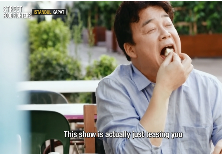 A screenshot from 'Street Food Fighter', Baek Jong-won taking a big bite of bread topped with kaymak