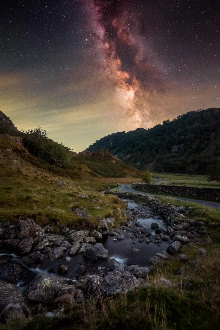 A scenic nighttime landscape showing the Milky Way glowing brilliantly above rolling hills, a winding road, and a rocky stream.
