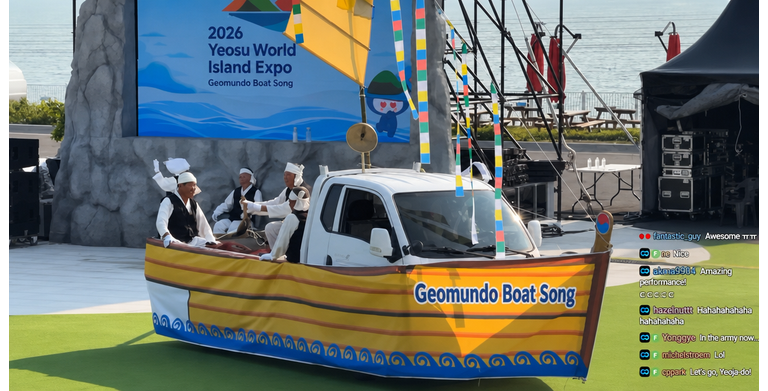 Performers sitting on a white truck decorated with a boat-shaped panel featuring the text "Geomundo Boat Song" on the grass in front of the Yeosu Island Expo stage.