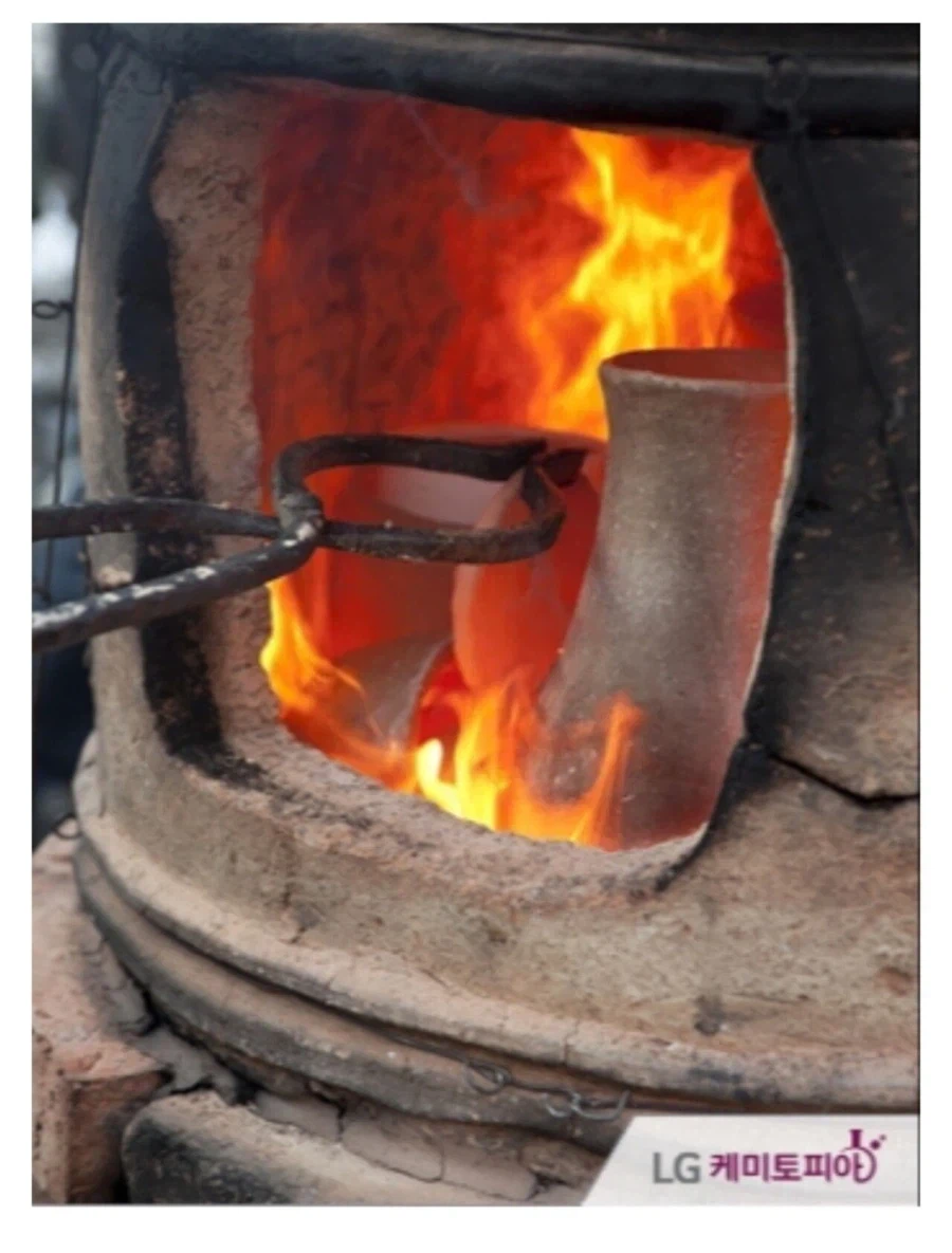 A glowing red ceramic piece being handled with metal tongs inside an earthen kiln with fierce red flames roaring