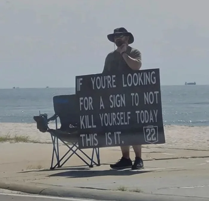 A man standing on a beach sidewalk next to a folding chair holding a large sign that reads: IF YOU'RE LOOKING FOR A SIGN TO NOT KILL YOURSELF TODAY THIS IS IT.