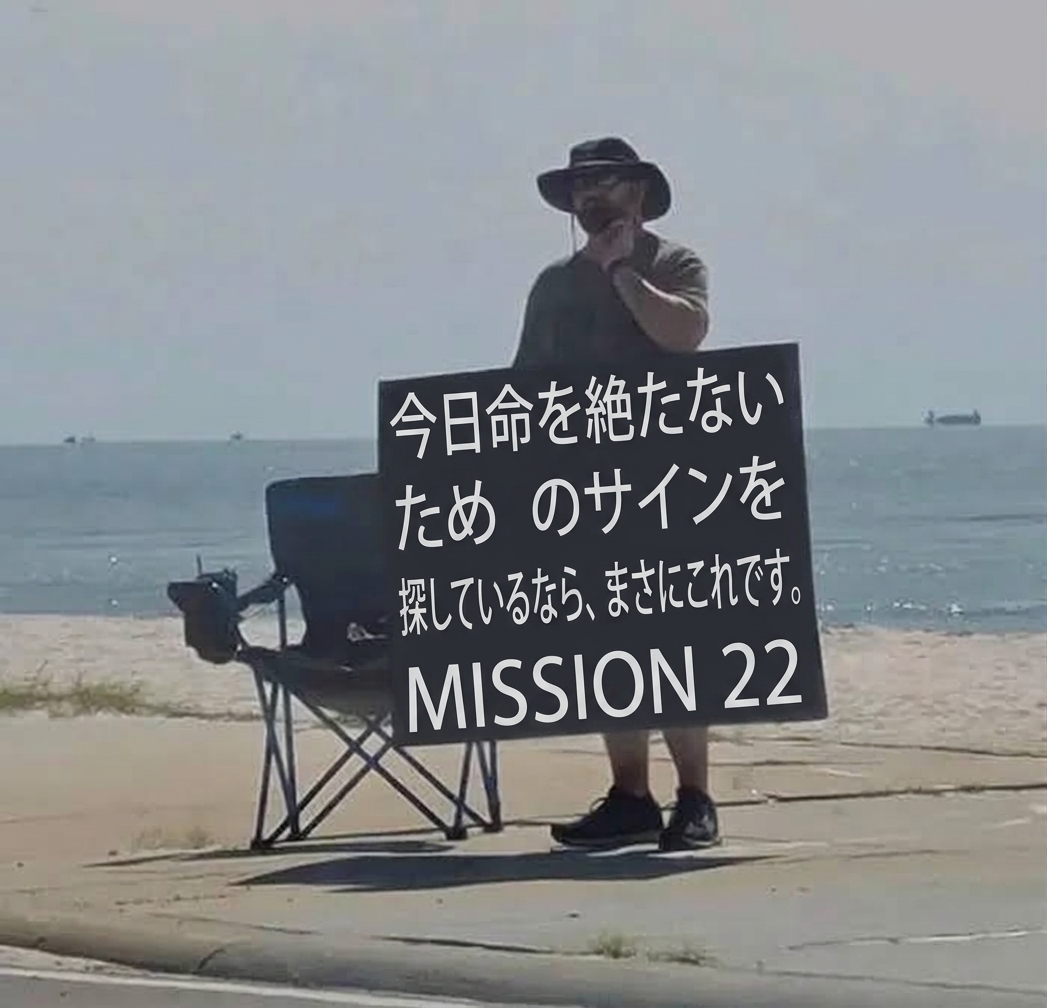 ビーチ沿いの歩道で折りたたみ椅子の横に立ち、「今日命を絶たないためのサインを探しているなら、まさにこれです」と書かれた大きな看板を掲げる男性。