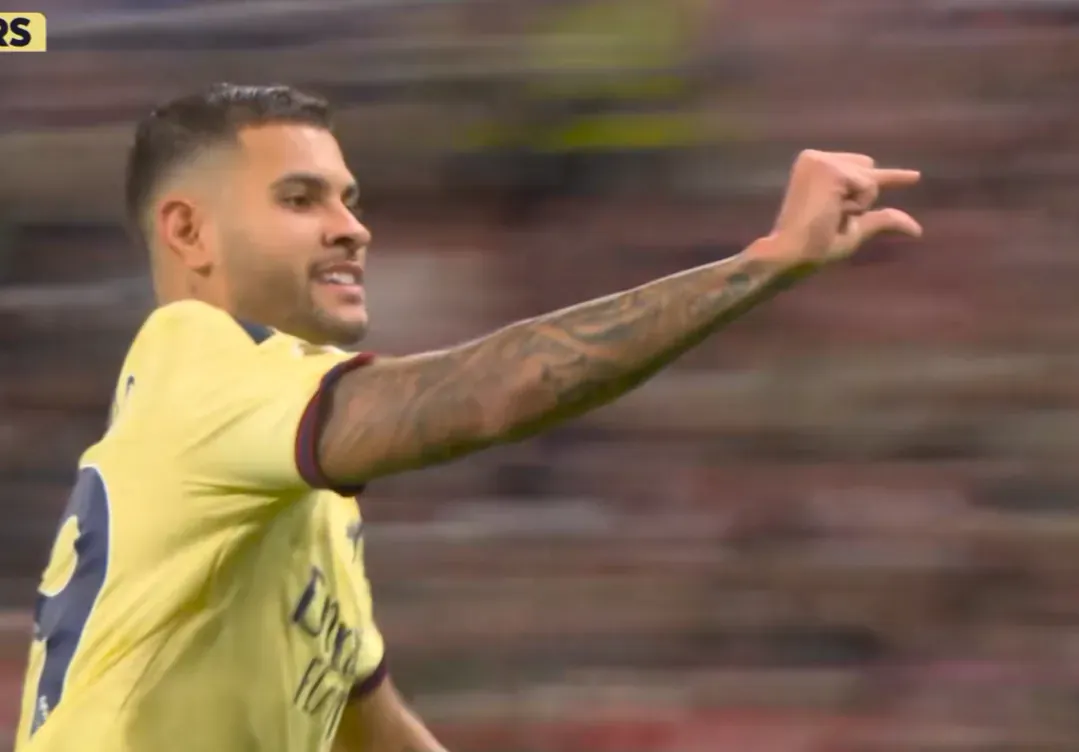 An Arsenal player in a yellow uniform extends an arm and makes a pinching gesture with the thumb and index finger.
