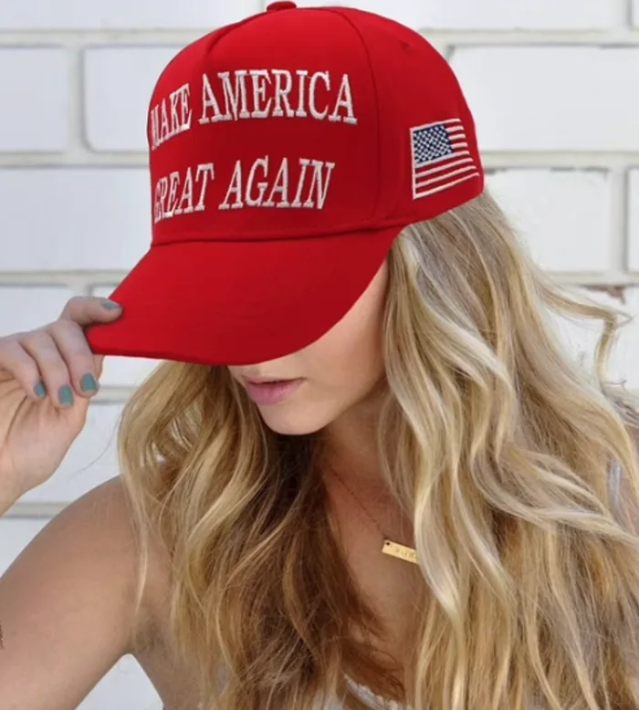 A blonde woman tilting down the brim of a red baseball cap embroidered with 'MAKE AMERICA GREAT AGAIN' and an American flag patch.