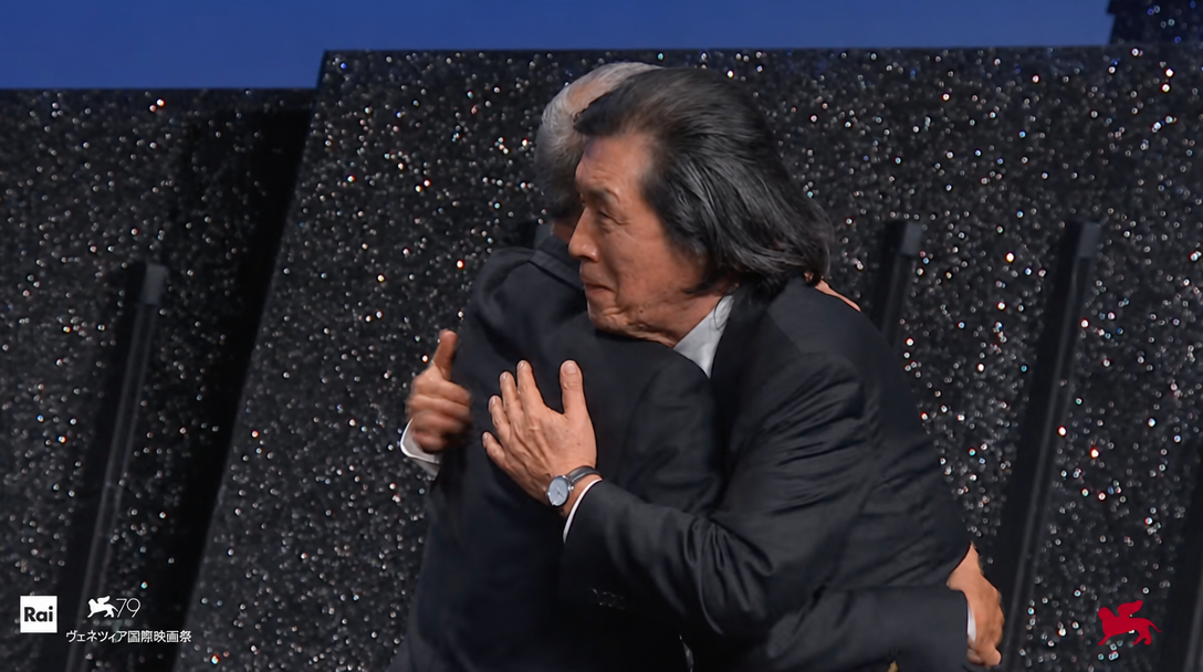 映画祭の授賞式のステージ上で、スーツを着た2人の男性が力強く抱き合っている様子。