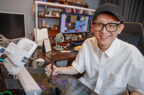 Webtoon artist Cho Seok wearing glasses and a black cap, smiling brightly with a stylus pen in his hand at his studio desk