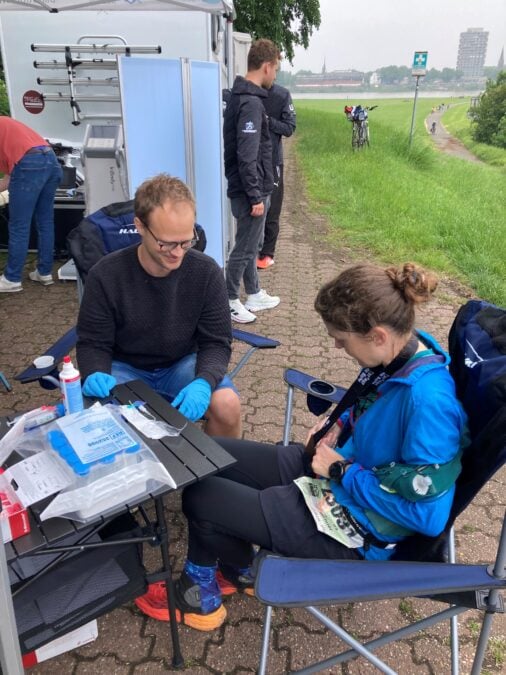 A researcher wearing blue gloves draws a blood sample from a female marathon participant in running attire seated at a makeshift table under an outdoor canopy.