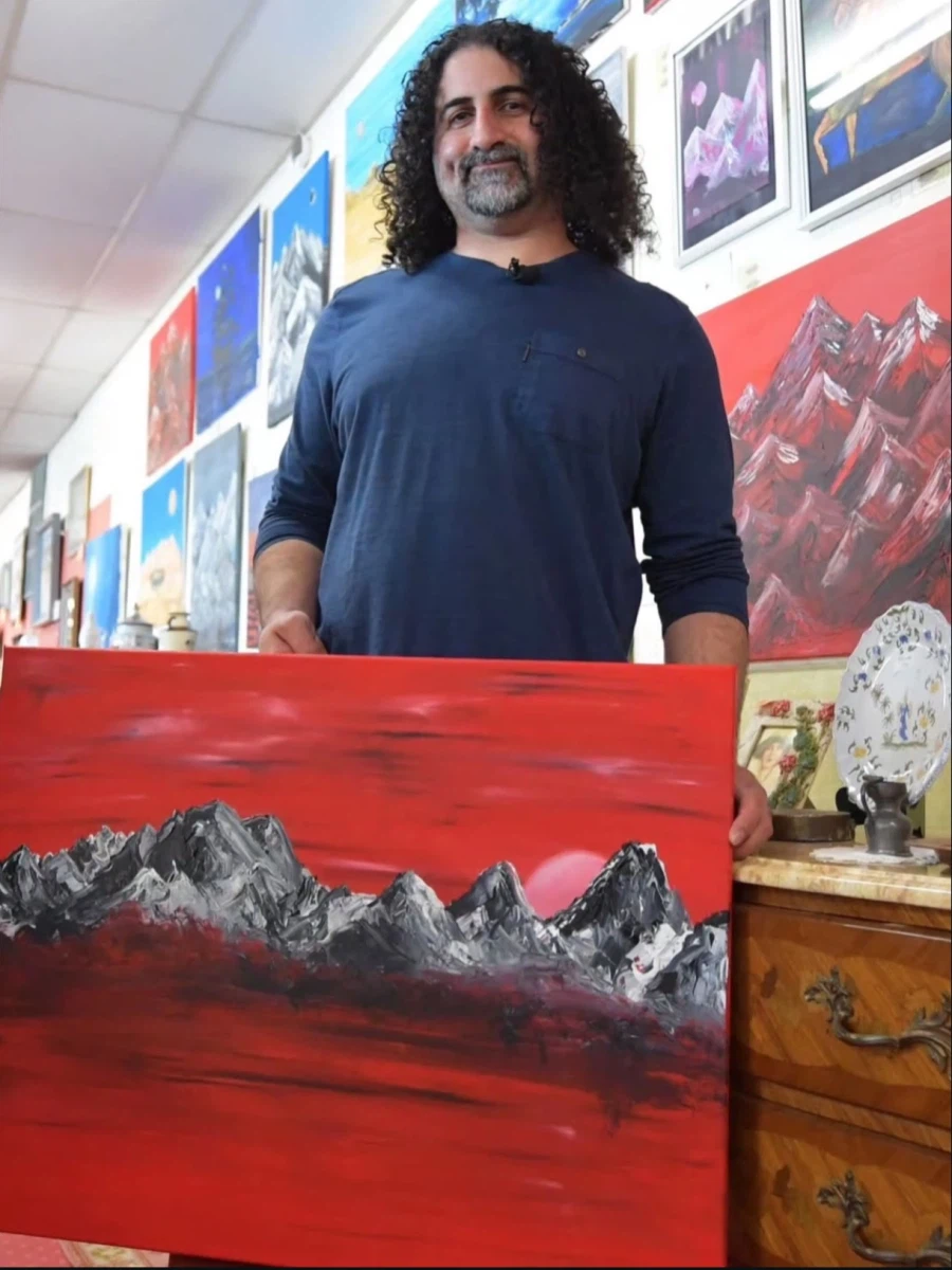 Omar bin Laden standing and holding his own painting of a mountain range on a red background