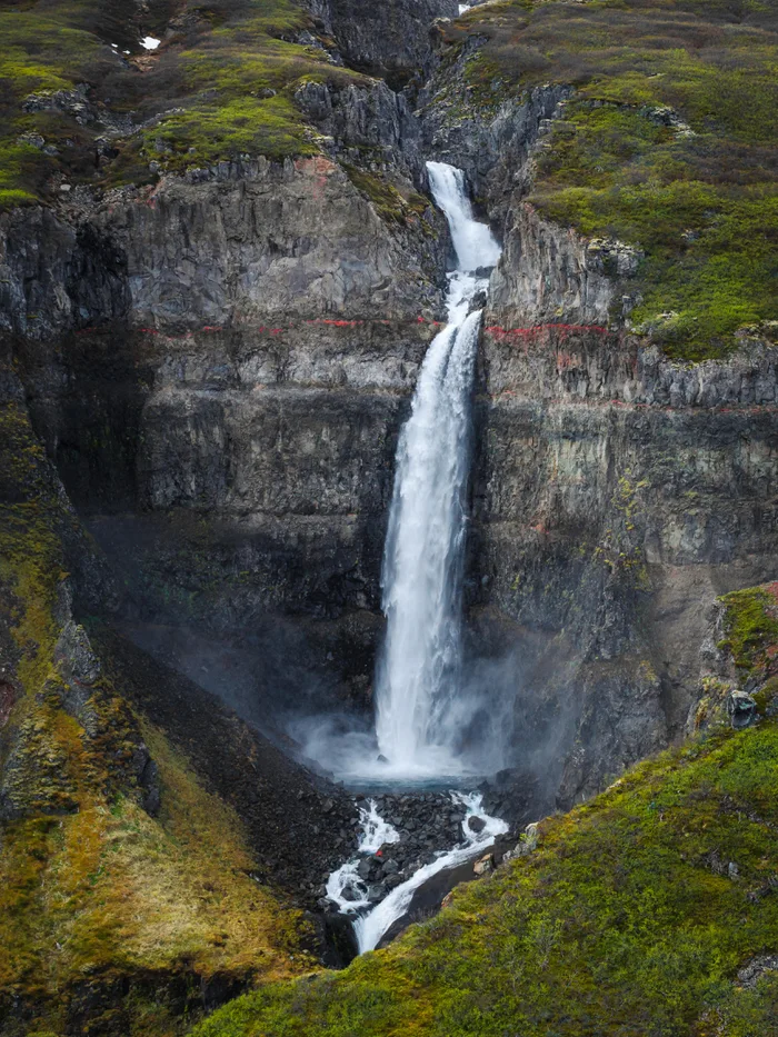 A tall, powerful waterfall flowing down a steep rocky cliffside with distinct reddish geological strata and mossy slopes.